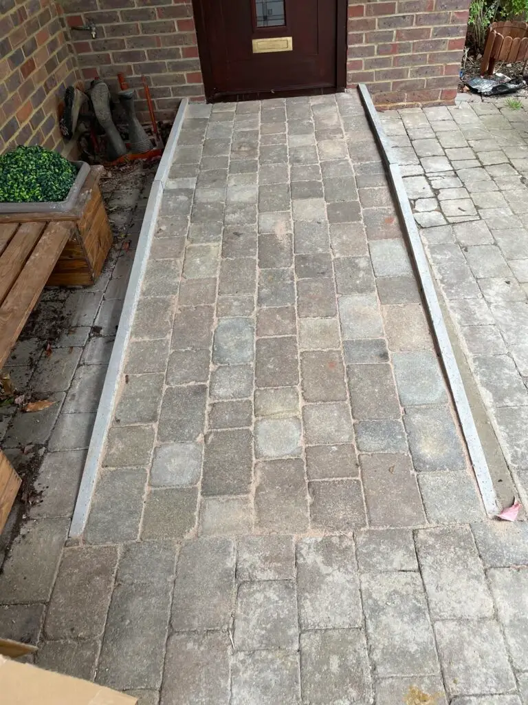 A stone paved wheelchair ramp leads up to a front door, bordered by two raised concrete edges and surrounded by a brick patio.
