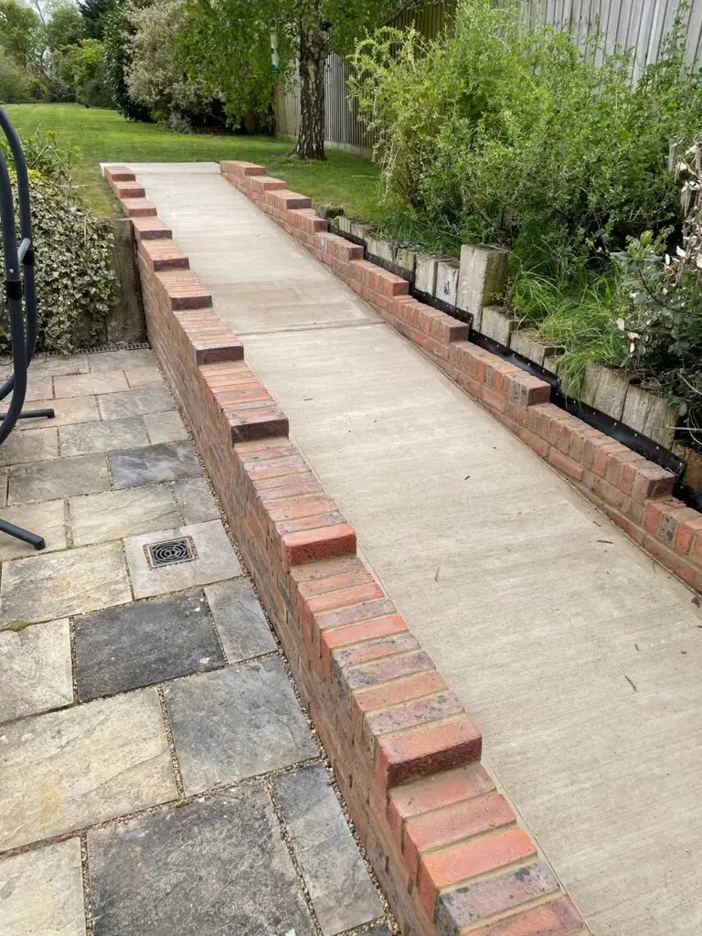 A paved outdoor walkway with low brick walls on each side leads from a stone patio to a grassy garden area, bordered by plants and trees.