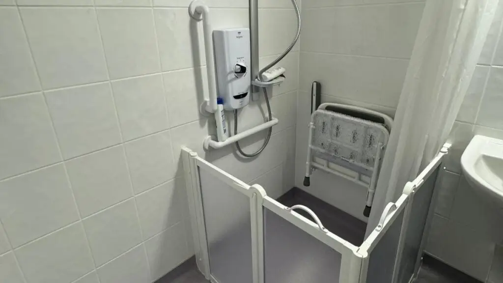 About us: Accessible shower area featuring a fold-down shower seat, grab bars, an electric shower unit, and a low-level folding splash guard enclosure.